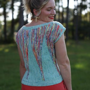 May include: A light blue knitted top with a scoop neck and short sleeves. The top features vertical stripes of red, yellow, and purple. The woman is wearing red pants and gold earrings.