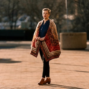 May include: A woman wearing a patterned shawl in shades of red, brown, and beige. The shawl is draped over a dark blue shirt and black leggings. She is wearing brown ankle boots. The background is a brick-paved area.