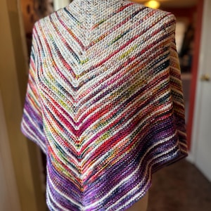 May include: A colorful, hand-knitted shawl with a V-neck design. The shawl features a vibrant array of colors, including purple, red, white, and yellow, arranged in diagonal stripes. The garment is displayed on a mannequin, showcasing its shape and texture.