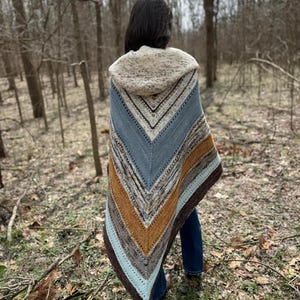 May include: A person wearing a hooded knitted shawl with a blue, brown and orange colour pattern. The shawl is worn over a pair of blue jeans. The person is standing in a wooded area.