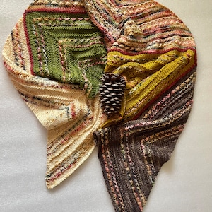 Könnte beinhalten: Ein dreieckiger gestrickter Schal in Braun-, Grün-, Gelb- und Cremetönen mit einer Kiefernzapfen auf der Oberseite.