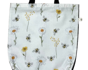 May include: White canvas tote bag with black handles and a repeating pattern of yellow daisies, green stems, and black and yellow bees.