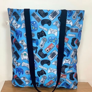 May include: A blue tote bag with black straps featuring a repeating pattern of video game controllers and consoles.