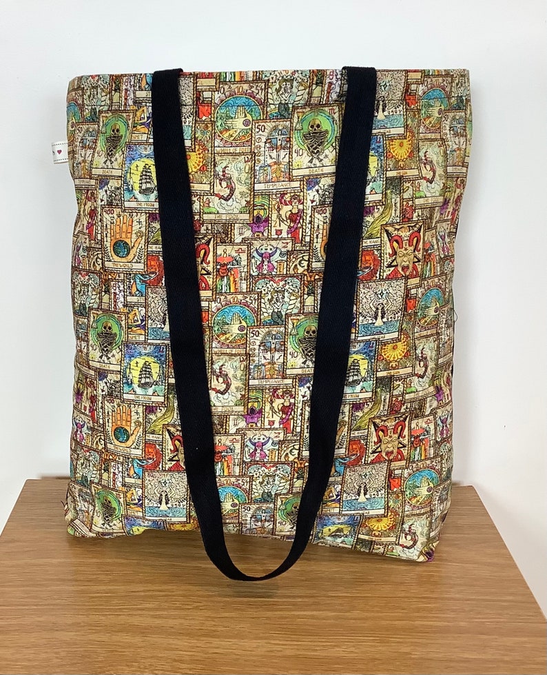 Op de afbeelding: Een grote tote bag met zwarte banden. De tas is gemaakt van beige stof met een kleurrijk patroon van tarotkaarten.