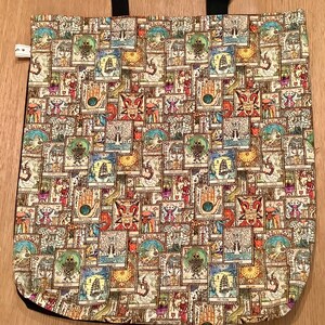 Op de afbeelding: Een beige tote bag met zwarte banden en een herhalend patroon van kleurrijke, ge&iuml;llustreerde tarotkaarten. De kaarten bevatten verschillende symbolen en afbeeldingen, waaronder schedels, schepen en handen.