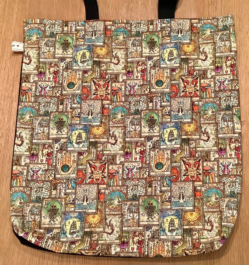 Op de afbeelding: Een beige tote bag met zwarte banden en een herhalend patroon van kleurrijke, ge&iuml;llustreerde tarotkaarten. De kaarten bevatten verschillende symbolen en afbeeldingen, waaronder schedels, schepen en handen.