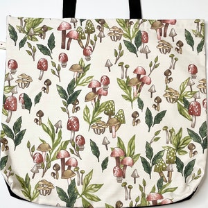 May include: A white canvas tote bag with black handles features a repeating pattern of red and brown mushrooms with green leaves on a white background.