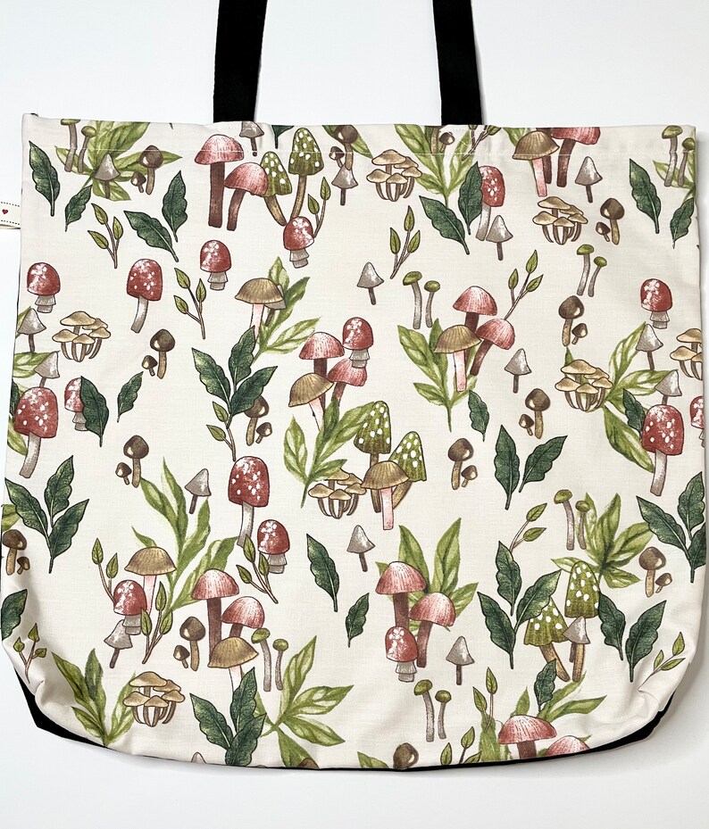 May include: A white canvas tote bag with black handles features a repeating pattern of red and brown mushrooms with green leaves on a white background.