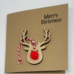 Op de afbeelding: Bruine kerstkaart met een houten rendierkop. Het rendier heeft een rode pomponneus en een rood-wit touwtje. De woorden "Merry Christmas" zijn donkerbruin gedrukt.
