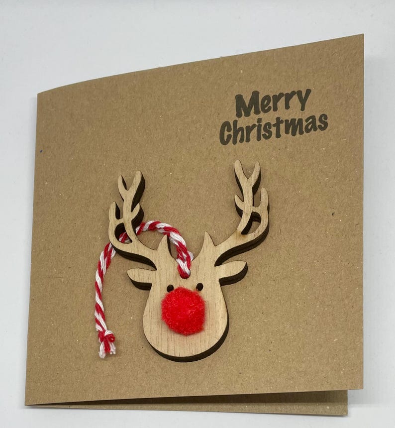 Op de afbeelding: Bruine kerstkaart met een houten rendierkop. Het rendier heeft een rode pomponneus en een rood-wit touwtje. De woorden "Merry Christmas" zijn donkerbruin gedrukt.