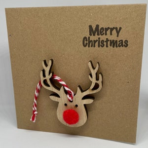 Op de afbeelding: Een vierkante, bruine kerstkaart met de woorden "Merry Christmas" in donkerbruin gedrukt. Een houten rendier met een rode neus en een rood-wit touwtje is aan de kaart bevestigd.