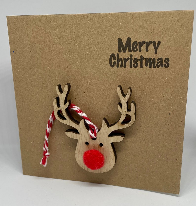 Op de afbeelding: Een vierkante, bruine kerstkaart met de woorden "Merry Christmas" in donkerbruin gedrukt. Een houten rendier met een rode neus en een rood-wit touwtje is aan de kaart bevestigd.