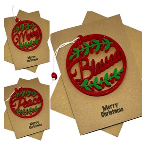 Op de afbeelding: Kerstversieringen met rode glitterletters en groene bladaccenten. De ornamenten zeggen "Merry", "Blessed" en "Peace". Elk ornament is bevestigd aan een kraftpapier kaart met de tekst "Merry Christmas".