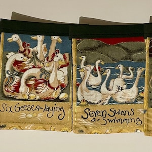 May include: A set of four fabric wall banners with a green border. Each banner features a different scene from the song "The Twelve Days of Christmas". The banners depict five gold rings, six geese a-laying, seven swans a-swimming, and eight maids a-milking.