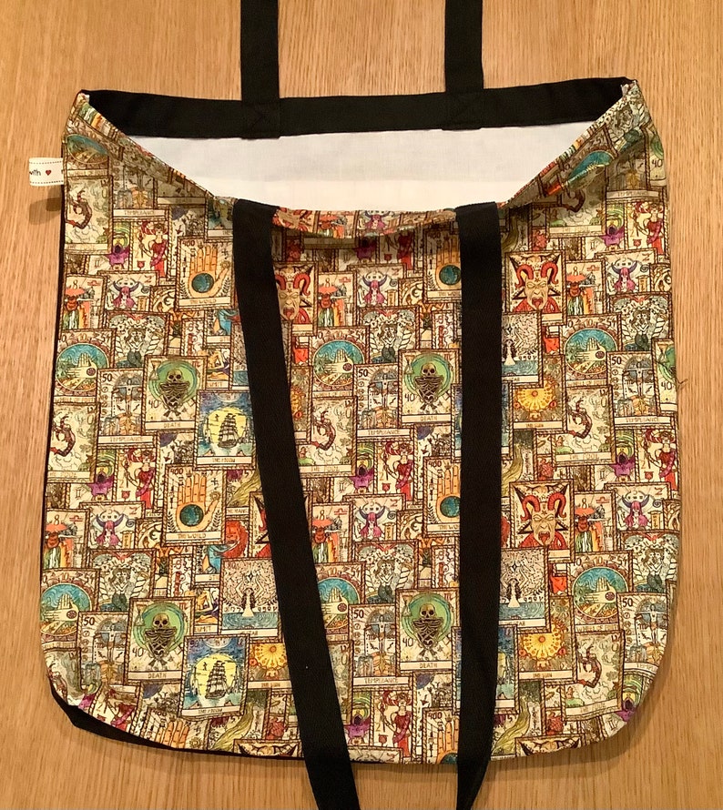 Op de afbeelding: Een beige tote bag met zwarte banden. De tas is gemaakt van canvas en heeft een patroon van kleurrijke illustraties.