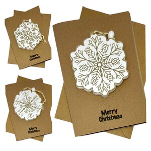 Op de afbeelding: Kerstkaarten van kraftpapier met een wit en goudkleurig sneeuwvlokornament. De kaarten hebben de tekst "Merry Christmas" in het zwart. De sneeuwvlokornamenten zijn gedetailleerd met een gouden ontwerp en een touwtje.