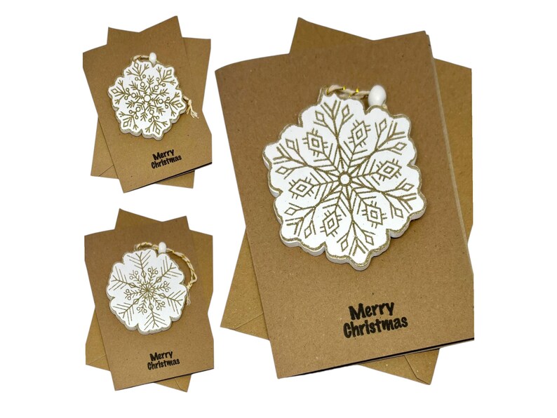 Op de afbeelding: Kerstkaarten van kraftpapier met een wit en goudkleurig sneeuwvlokornament. De kaarten hebben de tekst "Merry Christmas" in het zwart. De sneeuwvlokornamenten zijn gedetailleerd met een gouden ontwerp en een touwtje.