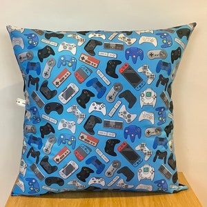 May include: A blue pillow with a pattern of various video game controllers and consoles. The controllers are in black, white, and red, and the consoles are in white, black, and gray.