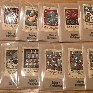 Op de afbeelding: Een verzameling kerstkaarten, elk afzonderlijk verpakt. De kaarten bevatten illustraties uit het lied "The Twelve Days of Christmas", met teksten als "Twee tortelduiven" en "Vrolijk Kerstfeest".