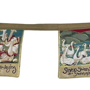 May include: A garland of fabric flags with illustrations and text from the song "The Twelve Days of Christmas". The flags are decorated with gold trim and feature images of five gold rings, six geese a-laying, seven swans a-swimming, and eight maids a-milking.