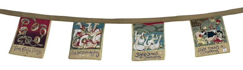 May include: A garland of fabric flags with illustrations and text from the song "The Twelve Days of Christmas". The flags are decorated with gold trim and feature images of five gold rings, six geese a-laying, seven swans a-swimming, and eight maids a-milking.
