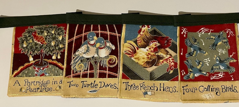 May include: A set of five fabric Christmas bunting flags with a red, green, and gold color scheme. Each flag features a different illustration from the Twelve Days of Christmas song: a partridge in a pear tree, two turtle doves, three French hens, four calling birds, and five golden rings. The flags are sewn together with a green ribbon.