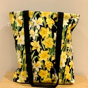 May include: A black tote bag with black straps and a floral print of yellow daffodils on a black background.
