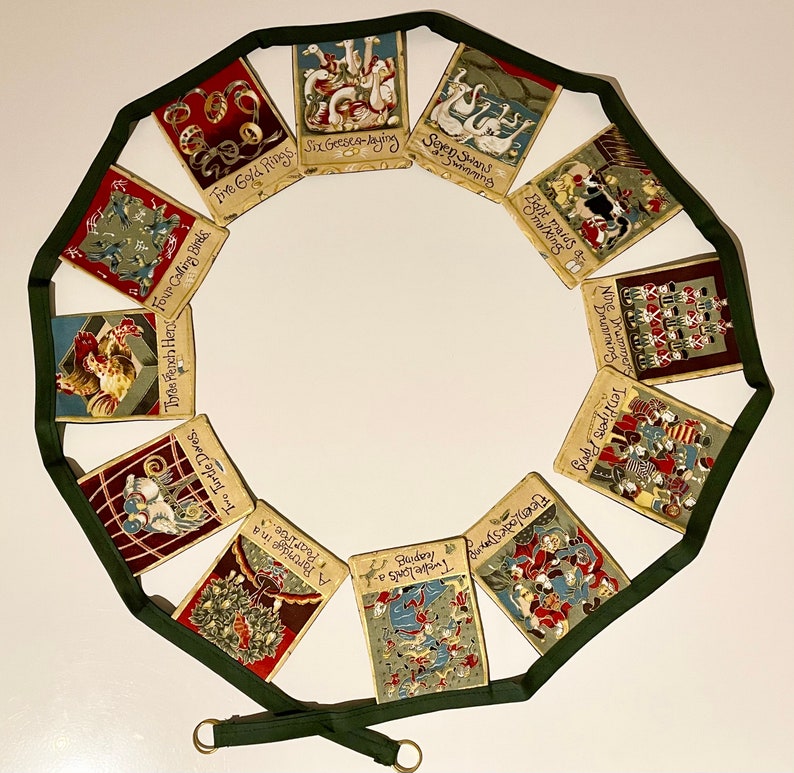 May include: A Christmas garland with 12 fabric ornaments featuring the 12 Days of Christmas song lyrics. Each ornament is decorated with a different illustration and the corresponding lyric. The garland is made of green ribbon and has a gold tassel at the end.