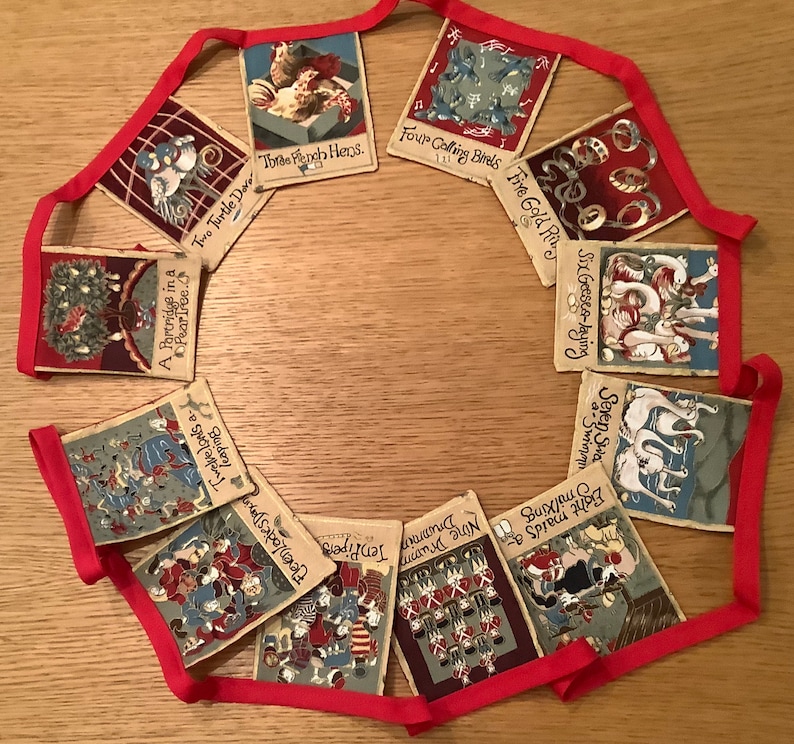May include: A red and white string of bunting with 12 flags. Each flag has a different illustration and text. The text on the flags includes: "A Partridge in a Pear Tree", "Two Turtle Doves", "Three French Hens", "Four Calling Birds", "Five Gold Rings", "Six Geese a-Laying", "Seven Swans a-Swimming", "Eight Maids a-Milking", "Nine Ladies Dancing", "Ten Lords a-Leaping", "Eleven Pipers Piping", and "Twelve Drummers Drumming".