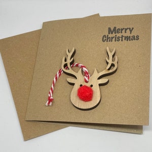 Op de afbeelding: Een kerstkaart van kraftpapier met een houten rendierkopornament. Het rendier heeft een rode pomponneus en een rood-wit touwtje. De woorden "Merry Christmas" zijn in het zwart op de kaart gedrukt.