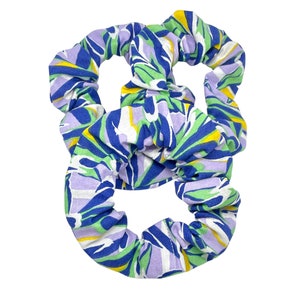 May include: Three scrunchies with a colorful abstract pattern. The fabric features a mix of blue, green, yellow, and lavender shapes on a white background. These hair accessories are designed to hold hair in place.