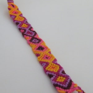 May include: A colourful friendship bracelet with a geometric pattern in shades of pink, orange, and purple.