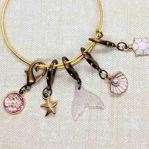 May include: Gold bangle bracelet with five charms: a pink iridescent charm, a starfish charm, a white mermaid tail charm, a white seashell charm with a pearl, and a white turtle charm.
