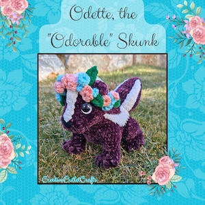Op de afbeelding: Een paars-witte gehaakte stinkdier met een bloemenkrans met roze, blauwe en groene bloemen. De stinkdier zit op een bed van groen gras. De tekst "Odette, de "Schattige" Stinkdier" staat boven de stinkdier.