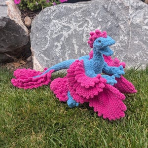 May include: A crocheted plush toy shaped like a dragon with a light blue body and vibrant hot pink frilled wings, a matching pink crest along the head, and a long, flowing pink-tipped tail, crafted from soft, textured yarn in a detailed design.