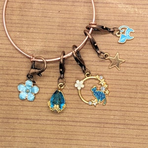 May include: A gold bangle bracelet with five charms: a blue flower, a teardrop-shaped blue gem, a gold circle with a blue cat and stars, a blue crescent moon, and a gold star.