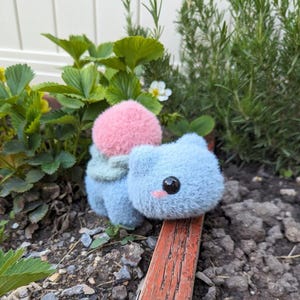 May include: A blue and pink plush toy resembling a Pokemon character. The toy has a large pink head and a smaller blue body with a green leaf-like collar.