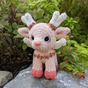 May include: A plush crochet creature featuring a light pink body, white antlers, and large black eyes. The figure is adorned with a decorative ribbon trim featuring small red roses around the neck and the base of the antlers, with darker pink hooves.
