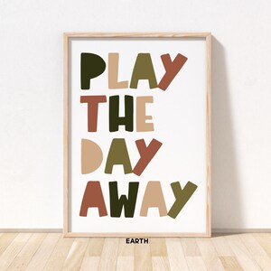 May include: A framed print with the text "PLAY THE DAY AWAY" in a colorful, playful font. The words are in shades of green, brown, and beige. The print is on a white background and has the word "EARTH" in black text at the bottom.