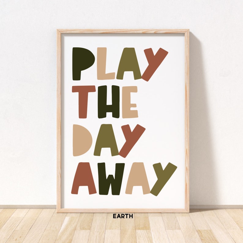 May include: A framed print with the text "PLAY THE DAY AWAY" in a colorful, playful font. The words are in shades of green, brown, and beige. The print is on a white background and has the word "EARTH" in black text at the bottom.