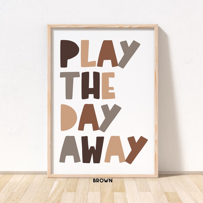 May include: A framed print with the text "PLAY THE DAY AWAY" in brown, tan, and gray letters. The text is in a playful, hand-drawn style. The frame is brown wood.