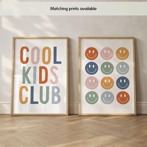 May include: Two framed wall art prints. The left print features the text "COOL KIDS CLUB" in multicolored block letters. The right print displays a grid of twelve colorful smiley face icons in shades of orange, pink, blue, and green.