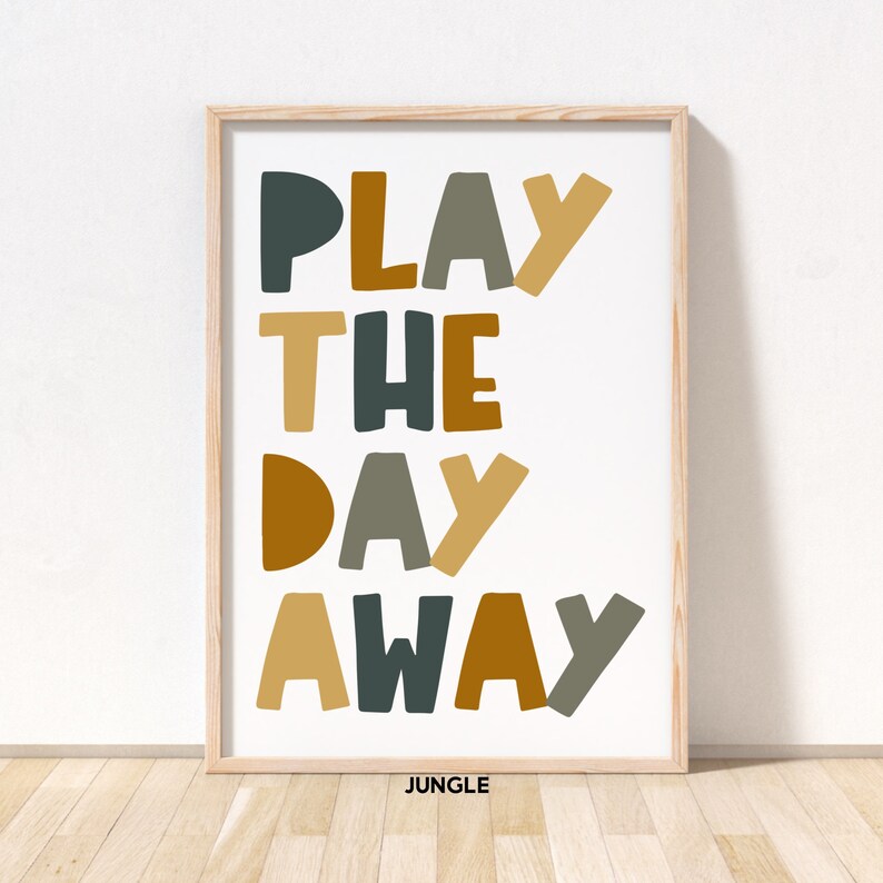 May include: A framed print with the text "PLAY THE DAY AWAY" in a colorful, playful font. The words are in shades of mustard yellow, brown, and teal. The print has a white background and a light wood frame. The word "JUNGLE" is printed in small letters at the bottom of the print.