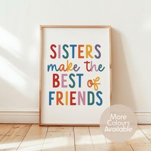 May include: A framed wall art print featuring the text "SISTERS make the BEST of FRIENDS" in a multi-coloured, rounded sans-serif font. The letters are displayed in shades of red, blue, yellow, purple, and green on a white background.