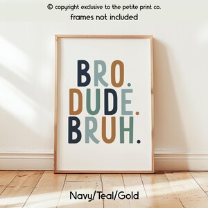 May include: A framed print with the words "BRO. DUDE. BRUH." in navy, teal, and gold colors. The print is on a white background and has a wooden frame. The text "Navy/Teal/Gold" is at the bottom. The text "copyright exclusive to the petite print co. frames not included" is at the top.