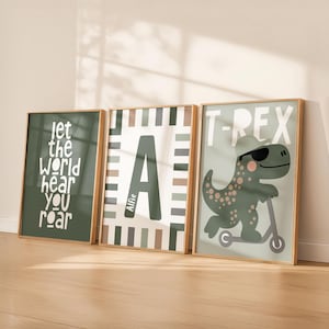 May include: Three framed wall art prints in light wood frames. The left print is dark green with white text reading 'let the world hear you roar'. The centre print features a large letter 'A' with the name 'Alfie' and a striped border. The right print shows a green dinosaur wearing sunglasses and a cap on a scooter with the text 'T-REX'.