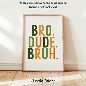May include: A framed art print with the words "BRO. DUDE. BRUH." in green and gold lettering. The phrase "Jungle Bright" is at the bottom. The print is in a wooden frame and leans against a white wall.