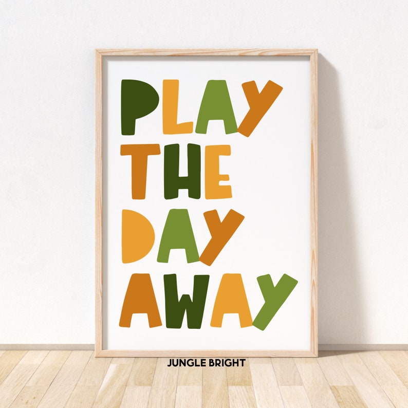 May include: A white framed print with the text "PLAY THE DAY AWAY" in colorful, overlapping letters. The letters are in shades of green, orange, and yellow. The text "JUNGLE BRIGHT" is printed in smaller letters below the main text.