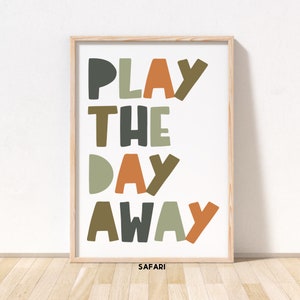 May include: A framed print with the text "PLAY THE DAY AWAY" in colorful, stylized letters. The letters are in shades of green, brown, orange, and gray. The print has a white background and a wooden frame. The text "SAFARI!" is printed in small letters at the bottom of the print.