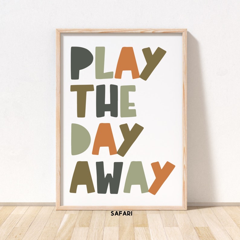 May include: A framed print with the text "PLAY THE DAY AWAY" in colorful, stylized letters. The letters are in shades of green, brown, orange, and gray. The print has a white background and a wooden frame. The text "SAFARI!" is printed in small letters at the bottom of the print.